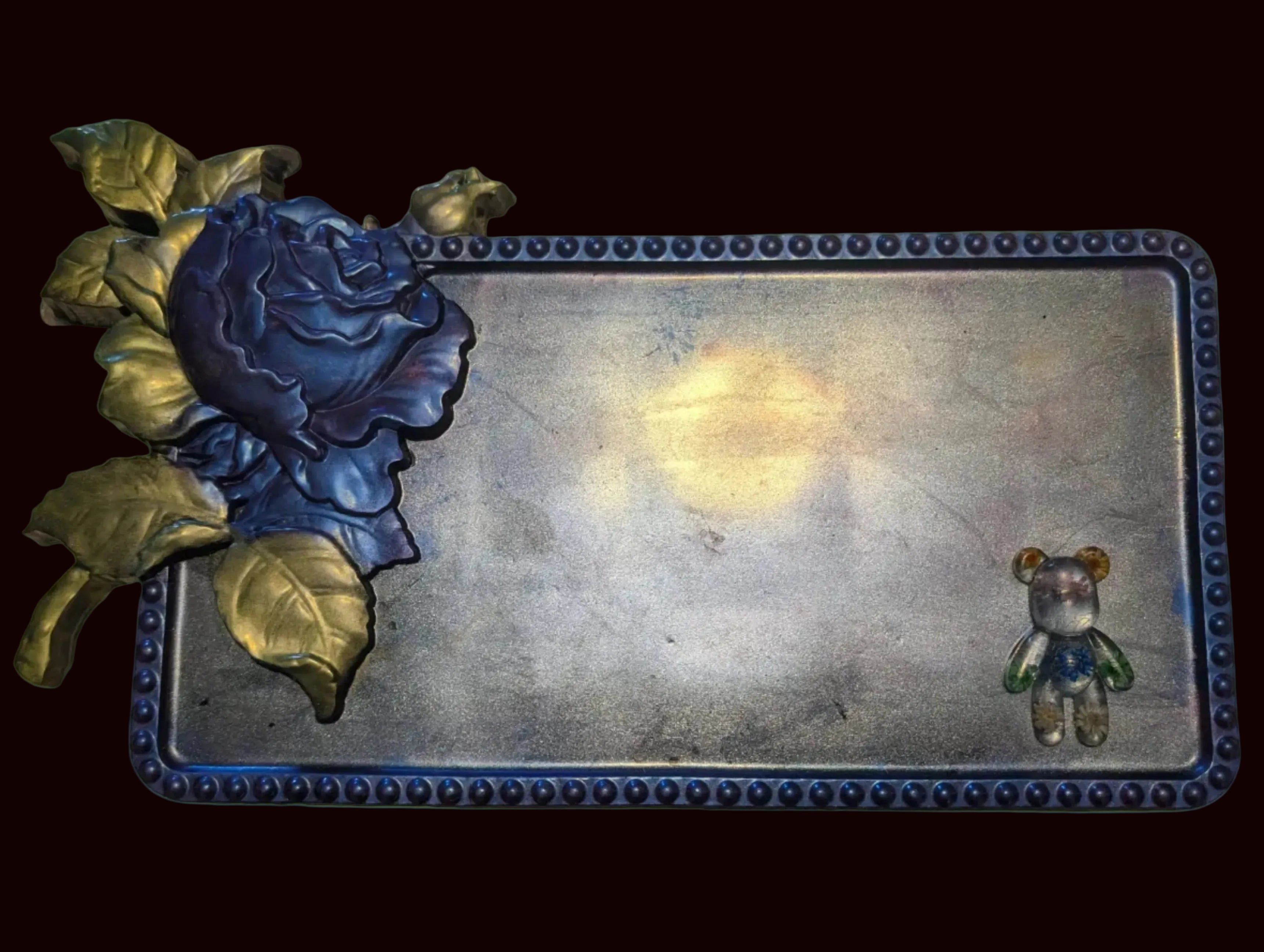 A rectangular decorative tray with a distressed silver-toned center, a dark blue beaded border, a large blue rose with gold leaves on the left, and a small teddy bear figure on the bottom right