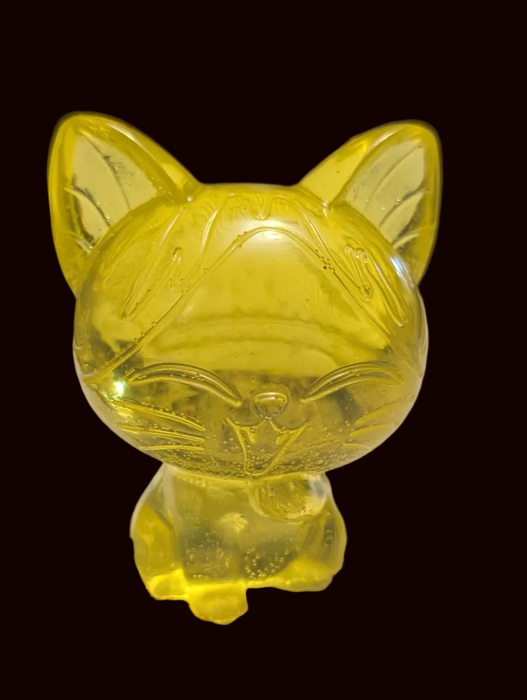 A small translucent yellow cat figurine or candle holder with pointed ears and a rounded body, standing on a white textured tabletop in a home setting with blurred background objects.