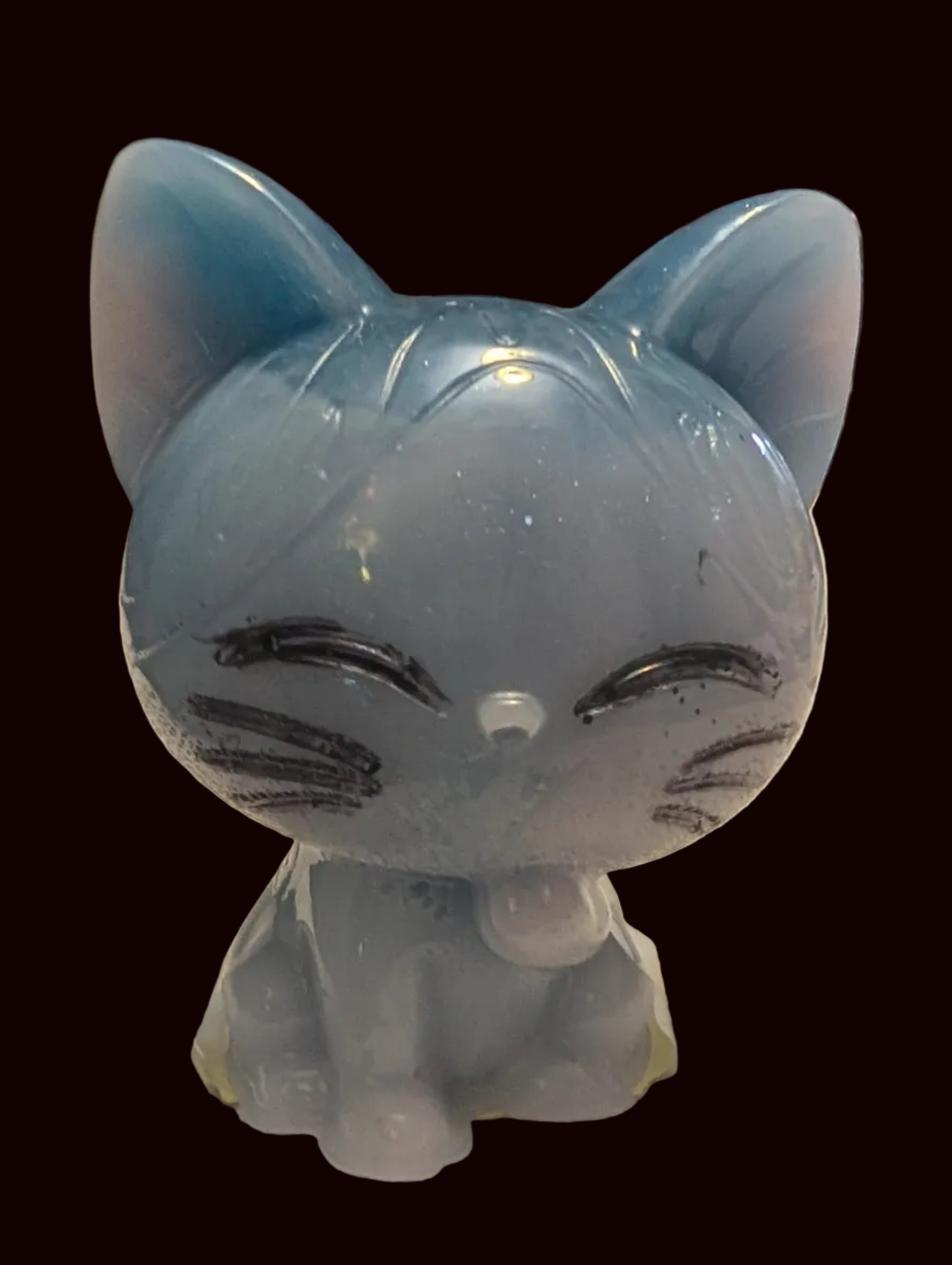 A small translucent blue cat figurine with closed eyes and whisker details, sitting on a white textured tablecloth in a home setting with a red clock in the background.
