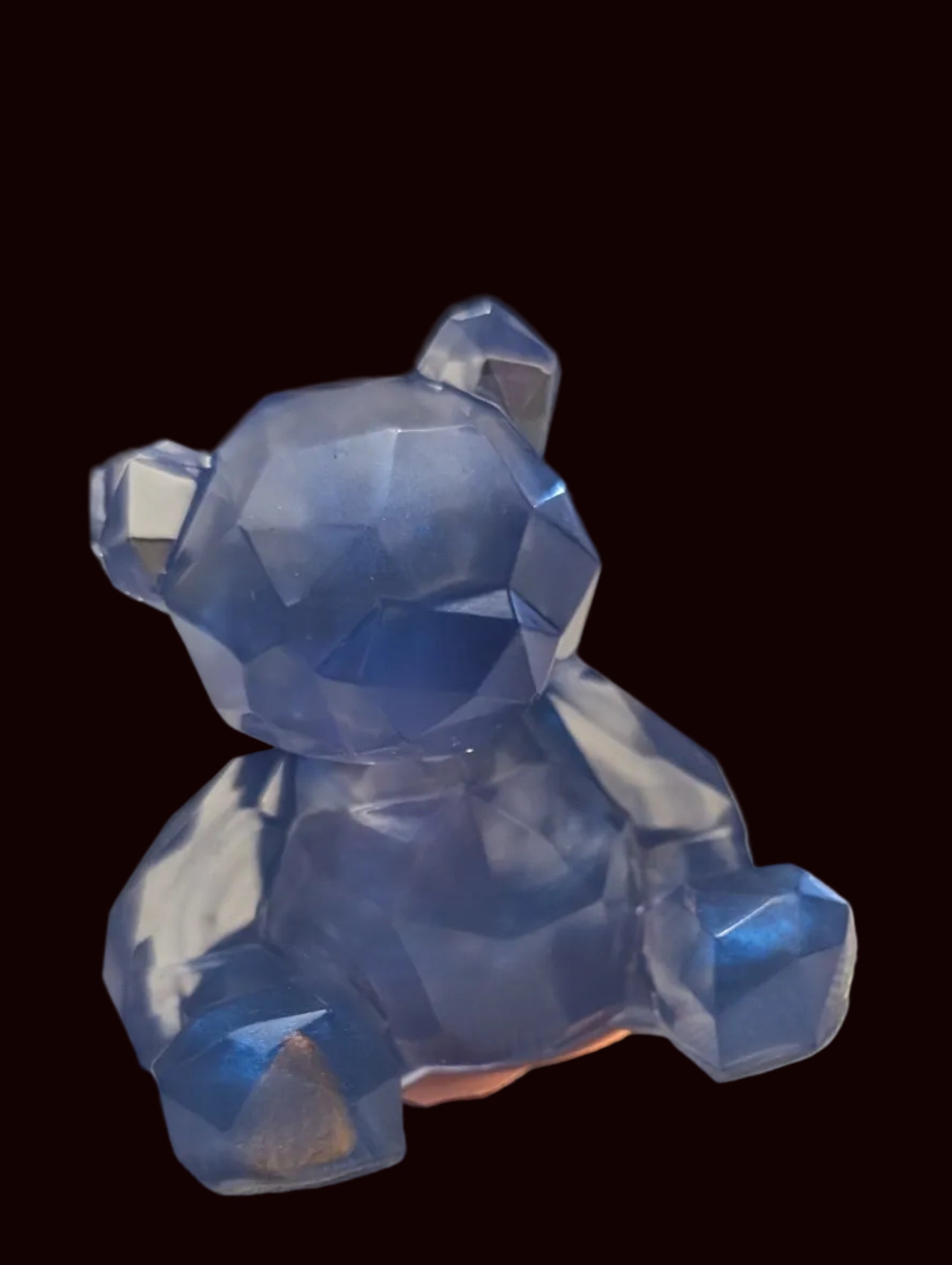 A small translucent blue faceted bear figurine sitting on a textured off-white surface, with blurred decorative objects in the background.