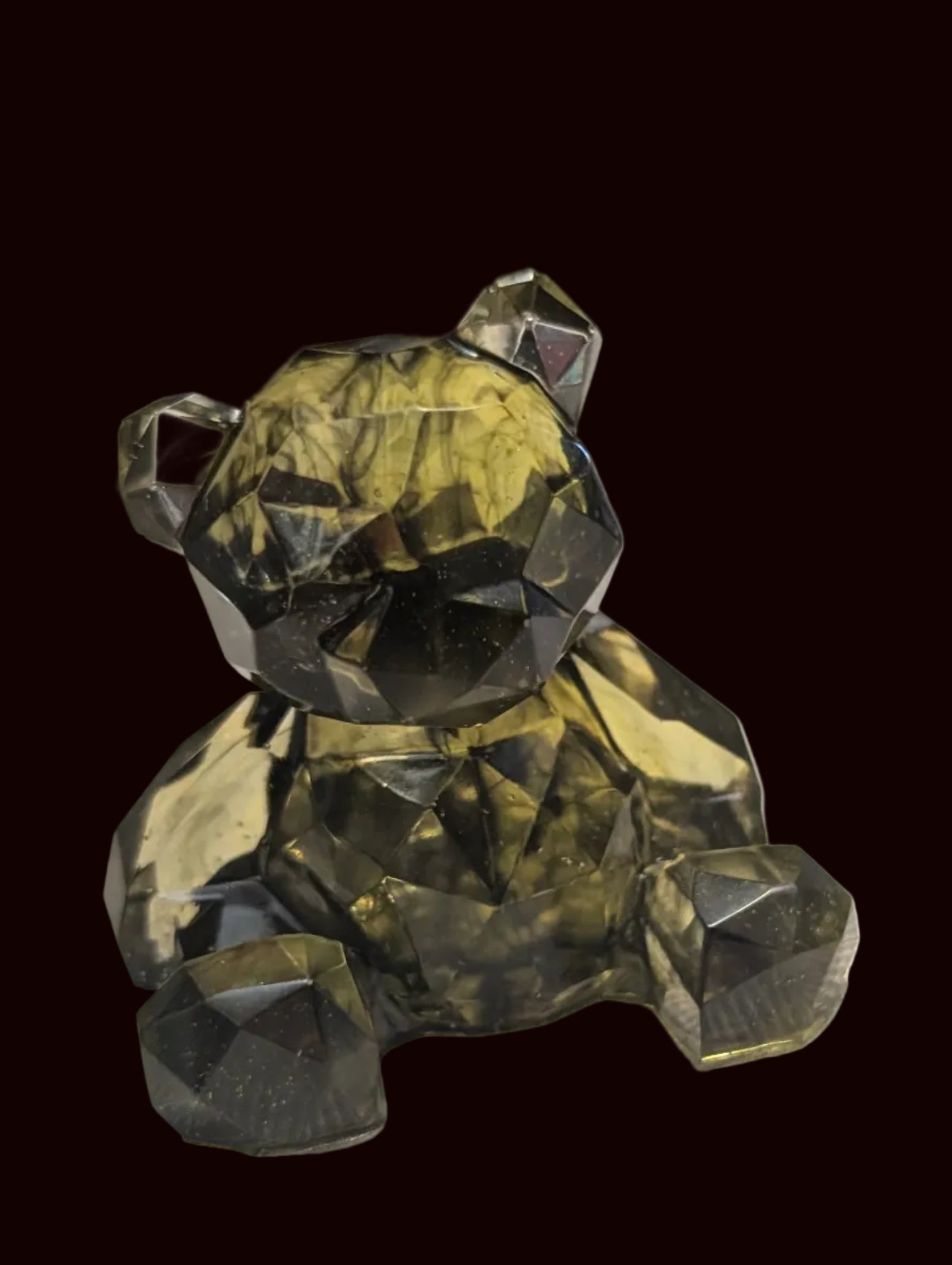 A faceted crystal miniature bear figurine in olive green and black, sitting on a textured white surface with a blurred background containing other decorative items.