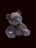 A small faceted crystal bear figurine with a dark purple body and a tiny red bow on its head, sitting on a white textured tablecloth with a blurred background.