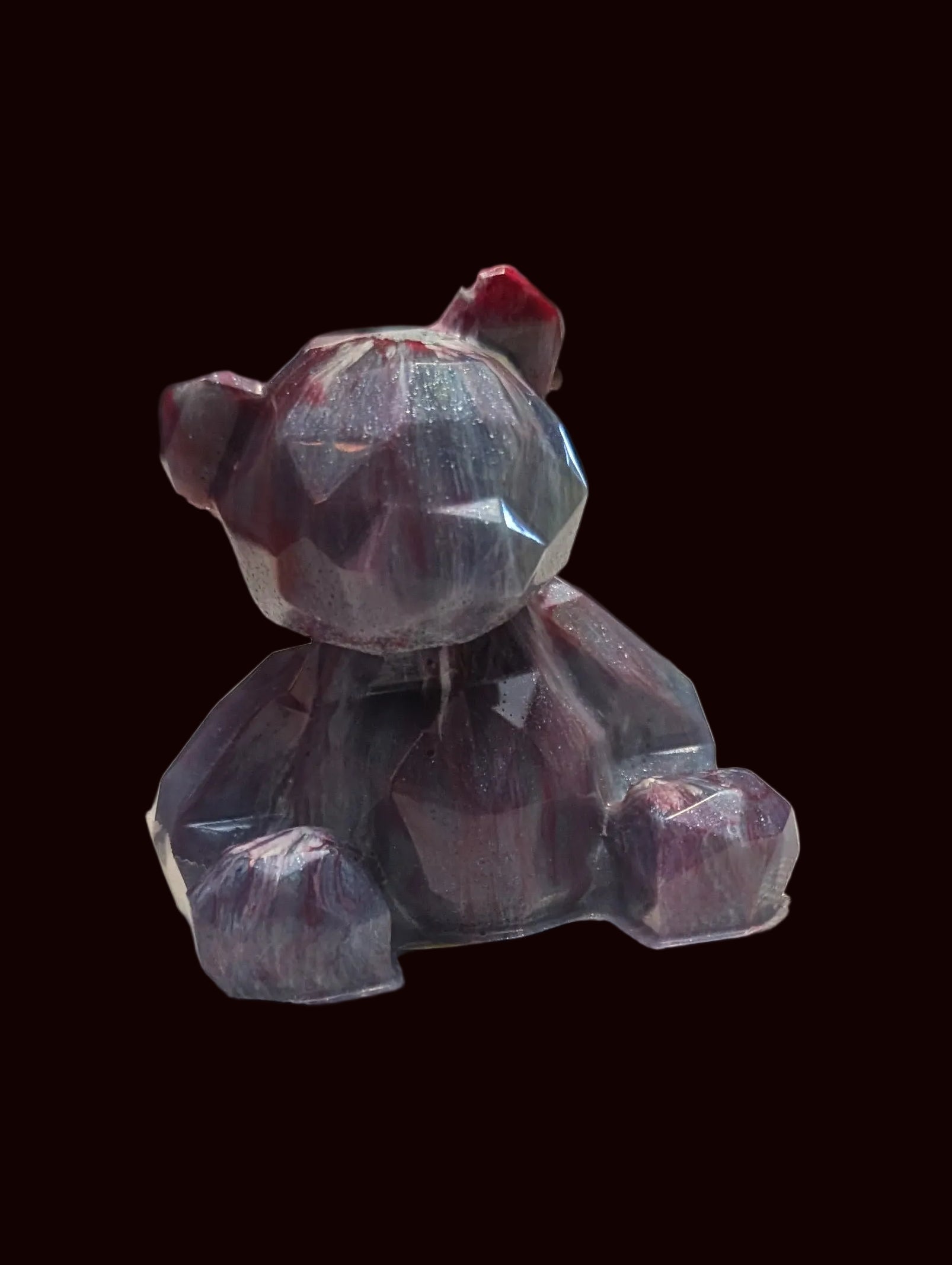 A small faceted crystal bear figurine with a dark purple body and a tiny red bow on its head, sitting on a white textured tablecloth with a blurred background.
