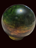 A polished iridescent crystal sphere with green, gold, and brown color shifts, sitting on a textured beige tablecloth with other colorful crystal figurines in the background.