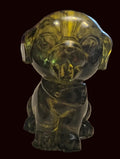 A small translucent green glass figurine of a dog sitting on a beige textured tablecloth, with a red wooden clock and other household items blurred in the background.