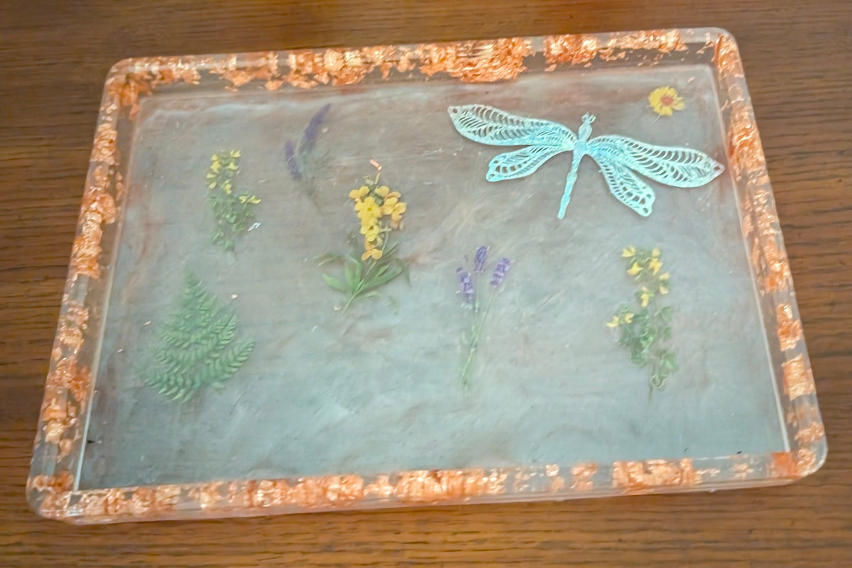 A square clear resin tray with a textured orange-brown border, featuring pressed botanical elements and a light blue dragonfly decoration, displayed on a dark wooden tabletop