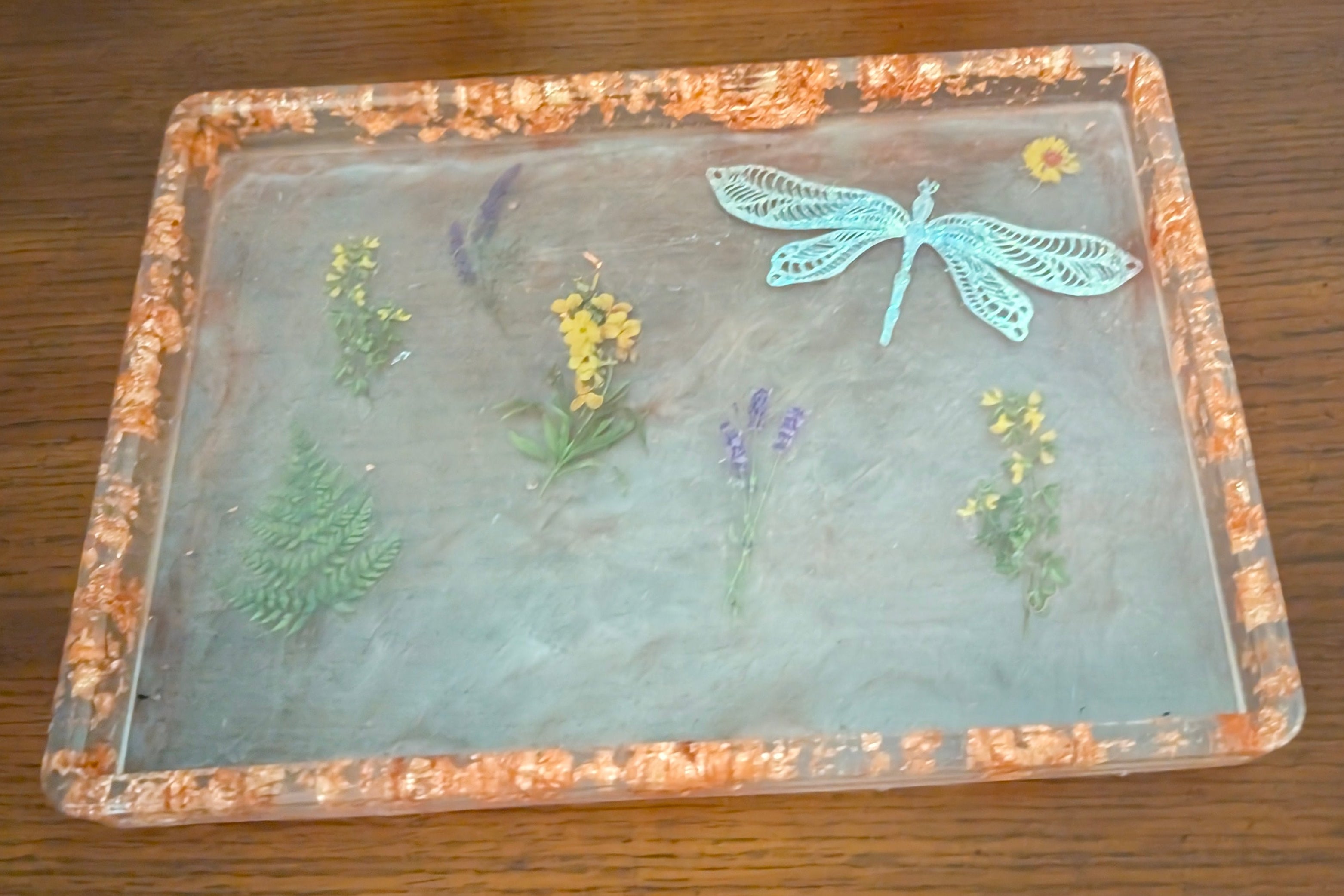 A square clear resin tray with a textured orange-brown border, featuring pressed botanical elements and a light blue dragonfly decoration, displayed on a dark wooden tabletop